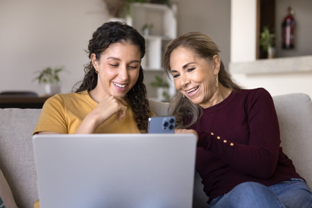 Two Latina women using laptop and smartphone. Mother and daughter choosing goods and gifts, calling to customer services to make purchase or booking hotels, buying tickets using marketplace platformの写真素材
