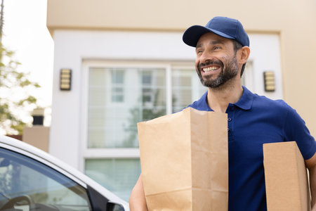 Happy middle aged courier man wearing blue uniform, carrying packages to car, enjoying logistic delivery job, looking away, smiling, laughing, walking from customers houseの写真素材