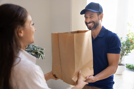 Cheerful positive delivery service manager in blue uniform cap giving paper packets with meal to...