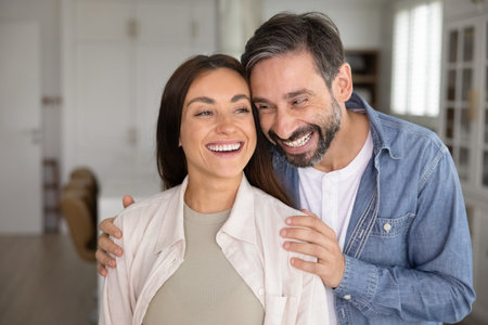 Happy attractive couple standing close together, hugging, looking away with toothy smiles, laughing, talking, discussing new house, apartment buying, enjoying home ownershipの写真素材