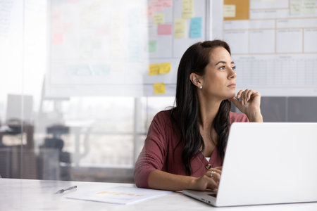 Thoughtful ambitious Hispanic business leader woman working at computer, looking away, touching...