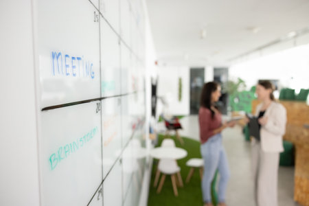Close up of scrum board in contemporary co-working space with two female business colleagues talking...