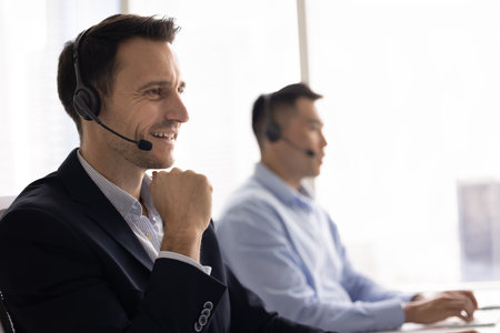 Positive Latin business professional man in headset speaking to client on audio call in contact center office, giving consultation, service, tech support, smiling, enjoying work communicationの写真素材