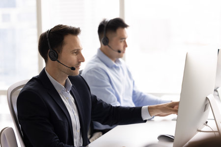Busy handsome contact center manager in wireless headset pointing at computer monitor at workplace, reviewing online content, talking on audio call, giving consultationの写真素材