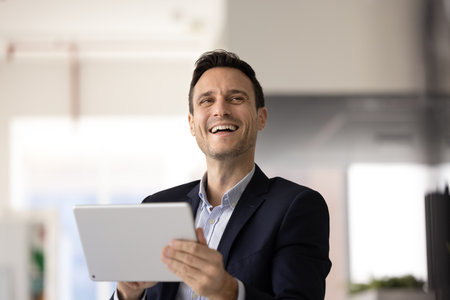 Happy joyful handsome business executive man using digital tablet for successful work result, typing on gadget, looking away, smiling, laughing, enjoying job success, thinking on company future visionの写真素材