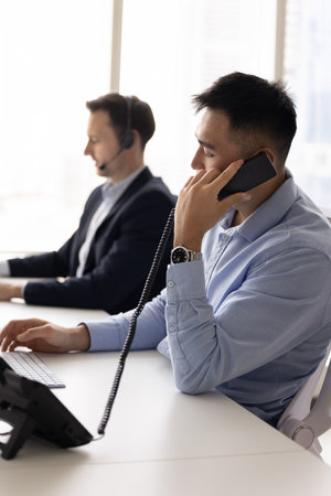 Asian contact center manager man talking to client on landline phone, holding handset, typing on...