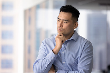 Serious thoughtful handsome Asian businessman touching chin, looking away, standing in office indoors, thinking on problem solving, complicated solution for project, softwareの写真素材