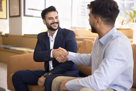 Two millennial 30s businessmen shaking hands, with joyful expression, feel satisfied with teamwork and partnership, met each other in office lobby, greeting, conclude successful meeting or agreementの写真素材