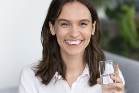 Happy pretty young adult girl drinking natural fresh water, holding glass, looking at camera with toothy smile, laughing, keeping healthy aqua balance, diet, lifestyle. Headshot home portraitの写真素材