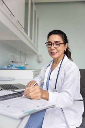 Positive focused practitioner doctor woman in glasses writing notes in paper medical records at workplace table, taking online professional training course, smiling, Vertical candid shotの写真素材