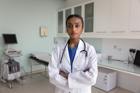 Confident serious young Black doctor woman posing in clinic ultrasonography room, looking at camera with hands folded in confidence gesture with ultrasound diagnosing computer in backgroundの写真素材