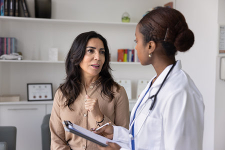 Mature Caucasian woman visiting young African female doctor for consultation, telling about healthcare, sickness symptoms. Physician filling medical form, listening to patient in clinic officeの写真素材