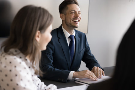 Laughing businessman in suit take part in briefing with teammates sit at conference table, share ideas, listen colleagues thoughts regarding joint project, engaged in productive, successful teamworkの写真素材