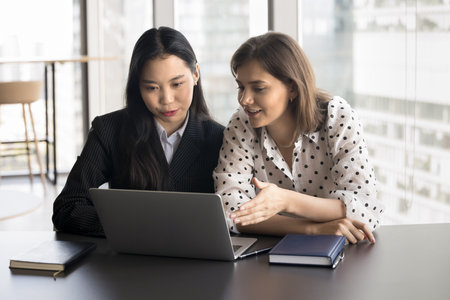 Young Asian and Caucasian women colleagues engaged in teamwork, preparing online presentation, working on joint project use laptop. Financial advisor consulting female bank client at meeting in officeの写真素材