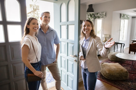Positive, real estate agent welcoming young married couple into house for sale or renting,...