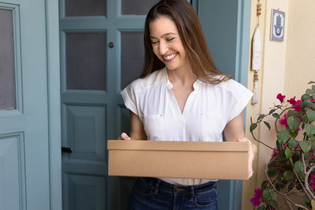 Beautiful smiling woman holding package standing on house porch feel excited of receiving long-awaited order or gift. Successful online shopping, customer satisfaction, doorstep delivery convenienceの写真素材