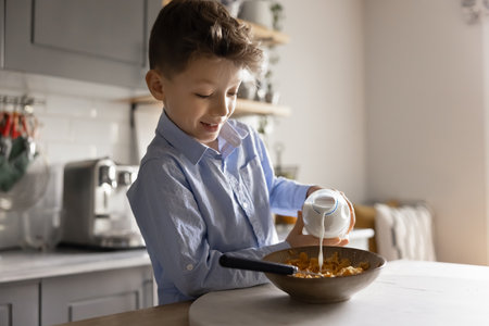 Cute little boy standing in modern kitchen in morning preparing independently breakfast before school, pouring cow or vegetal milk from bottle into his bowl full of sugar-free cereal. Balanced foodの写真素材