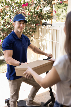 Smiling male courier in uniform on electric scooter delivering parcel box to female recipient...