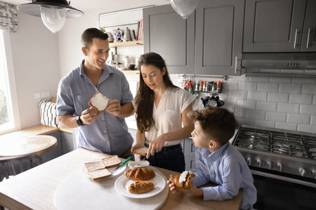 Family members engaged in preparing traditional breakfast, sharing morning moments together in modern kitchen. Young parents and little son gathered in cozy kitchen, cooking, butter bread, enjoy talkの写真素材