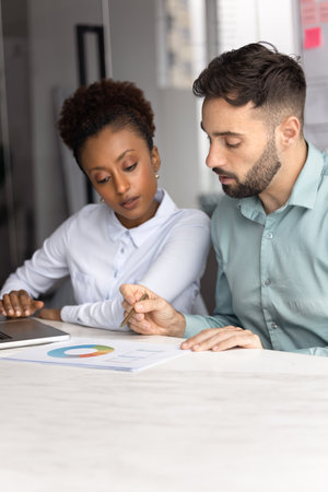 Two young diverse managers analyzing marketing report at office table, discussing diagram in paper document, working on project plan, sales growth strategy. Vertical shotの写真素材