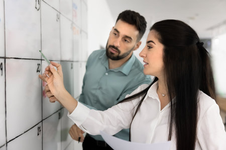 Two male and female Latin business colleagues using scrum management method for teamwork, working at Kanban board, writing in table cells on whiteboard, discussing business project planの写真素材