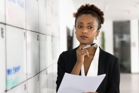 Pensive young Black business project manager woman thinking on tasking, scheduling at scrum table on Kanban board, touching chin, looking away, holding paper report, making decisionの写真素材