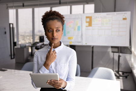 Serious young African American businesswoman holding tablet, thinking on online project electronic report, standing with digital gadget in office alone, looking away, touching chin, making decisionの写真素材