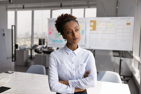 Serious young African American businesswoman standing with hands crossed in office workspace, looking away, thinking on business challenge, problem, solutions, planning project managementの写真素材