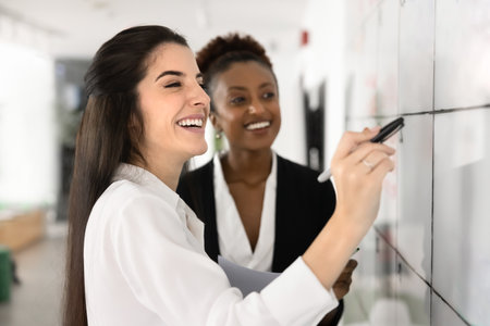 Cheerful Hispanic and African female coworkers discussing teamwork at Kanban board, working on business plan scheduling, tasking, enjoying teamwork, smiling, laughing, writing on whiteboardの写真素材