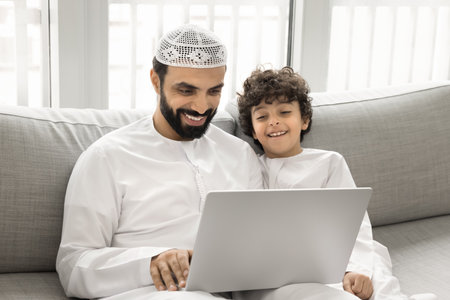 Cute little 7s boy and his young 30s dad dressed in white dishdasha using laptop, resting seated on sofa, watching comedy movie, or funny educational content, enjoy electronic shopping via marketplaceの写真素材