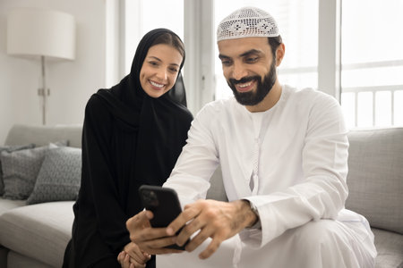 Arabian couple dressed in traditional dress, sit on sofa using smartphone, spend time on internet, get sell-out offer, watch video, viewing photos, engaging in online activity with joyful expressionの写真素材
