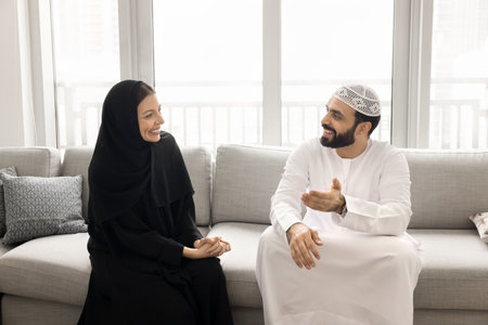 Happy couple sit on sofa, engaged in pleasant conversation, man dressed in traditional thobe lead talk with woman, wear black hijab, listens attentively. Understanding, cultural values, familial bondsの写真素材