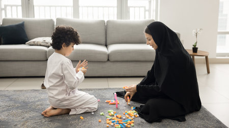 Middle Eastern mother dressed in black abaya playing blocks on floor with little son wearing white kandura, enjoy playtime in modern living room. Warmth, family bonding, quality time of mom and kidの写真素材