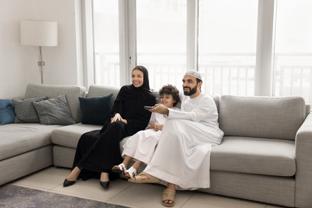 Young Middle Eastern family with little 7s son dressed in traditional Muslim attire watching smart TV seated on couch in modern living room, selecting movie or cartoons, enjoy favourite sitcom or showの写真素材