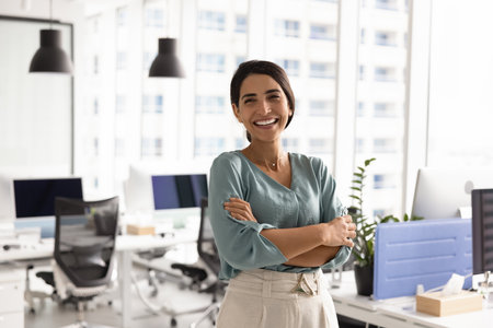 Happy successful female Latin businesswoman posing in office workspace alone, looking at camera, smiling, standing with arms crossed. Confident cheerful business leader woman portraitの写真素材
