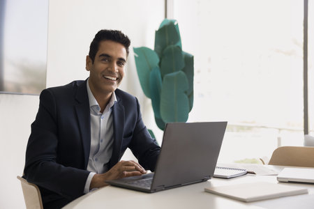 Portrait of mature Indian businessman sit at desk working on laptop, look at camera. Office employee dressed in suit managing business tasks on wireless computer, e-mailing, using business softwareの写真素材