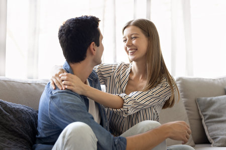 Young couple hugging, sitting close together on couch, smiling, looking into each other with...
