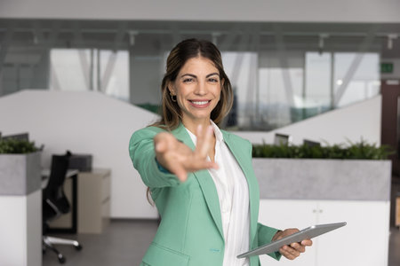 Positive beautiful Latin businesswoman making invitation hand gesture, inviting to join company team, welcoming for cooperation, partnership, offering professional support, consultationの写真素材