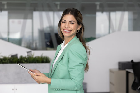 Cheerful beautiful Hispanic CEO woman holding digital gadget, looking at camera, smiling, standing in office with tablet. Latin female financial director, accountant, auditor, businesswoman portraitの写真素材