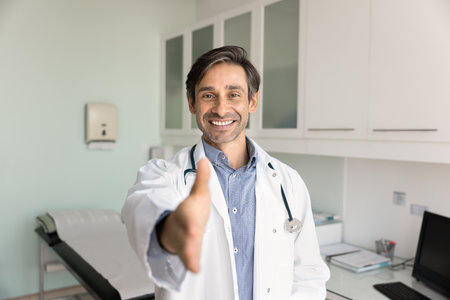 Portrait of middle-aged cardiologist in coat looks at camera, extending his arm for handshake, welcoming gesture, greeting clinic patient with positive expression. Trust, professionalism in healthcareの写真素材