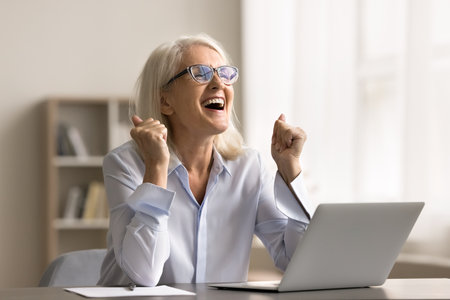 Success celebration. Portrait overjoyed excited elderly business lady executive scream yes great sitting by notebook at office desk get financial reward for perfect job loan approval marketing offerの写真素材