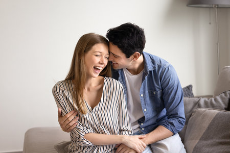 Couple sit on sofa, laughing, sharing joyful and intimate moments, exuding happiness and warmth. Loving man embracing his beloved woman, seated on couch at home. Romantic relations, holidays togetherの写真素材