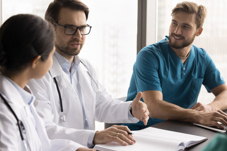 Collaboration in medicine. Confident young male head doctor lead round table briefing with diverse team of medical professionals discuss patients cases based on paper register offer treatment strategyの写真素材