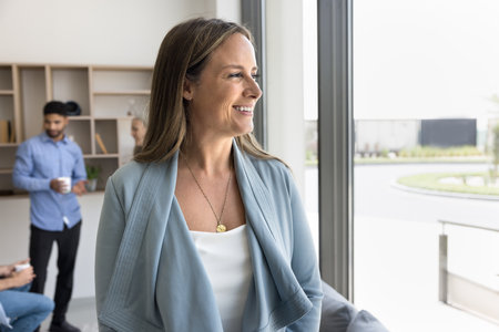 Successful businesswoman. Portrait happy beautiful mid aged lady business coach trainer leader stand by window at office workplace look away with confident smile proud of guiding team with experienceの写真素材