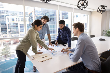 Indian professional woman expressing opinion, share solution, developing marketing strategy with diverse teammates gathered around conference table with heap of charts and graphs papers. Forecastingの写真素材