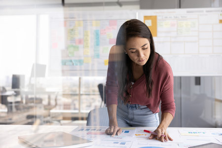 Developing project. Busy young woman business professional data analyst bend over desk with paper documents review reports read check statistic information. View through glass office wall. Copy spaceの写真素材
