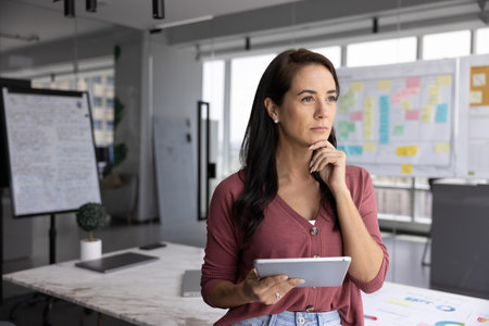 Creative strategy and online solutions. Pensive young lady stand in agile office space filled with visual planning tools holding digital pad consider work process organization reflect on business planの写真素材