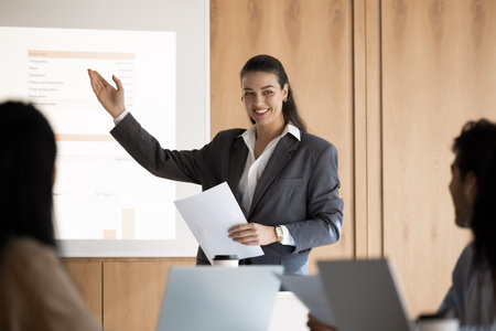Young 30s European business woman, female trainer or corporate coach presenting visual data, pointing on projected slide displaying analytical and financial information to diverse seminar participantsの写真素材