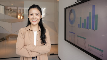 Professional confidence, leadership, and business intelligence. Portrait of Asian female marketing analyst or strategist standing in office next to screen displaying visual data with graphs and chartsの写真素材