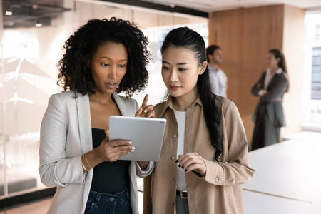 Serious African and Vietnamese women teammates using tablet in office, look at device screen with focused expression, collaborating on joint online task or project, sharing ideas or editing documentの写真素材